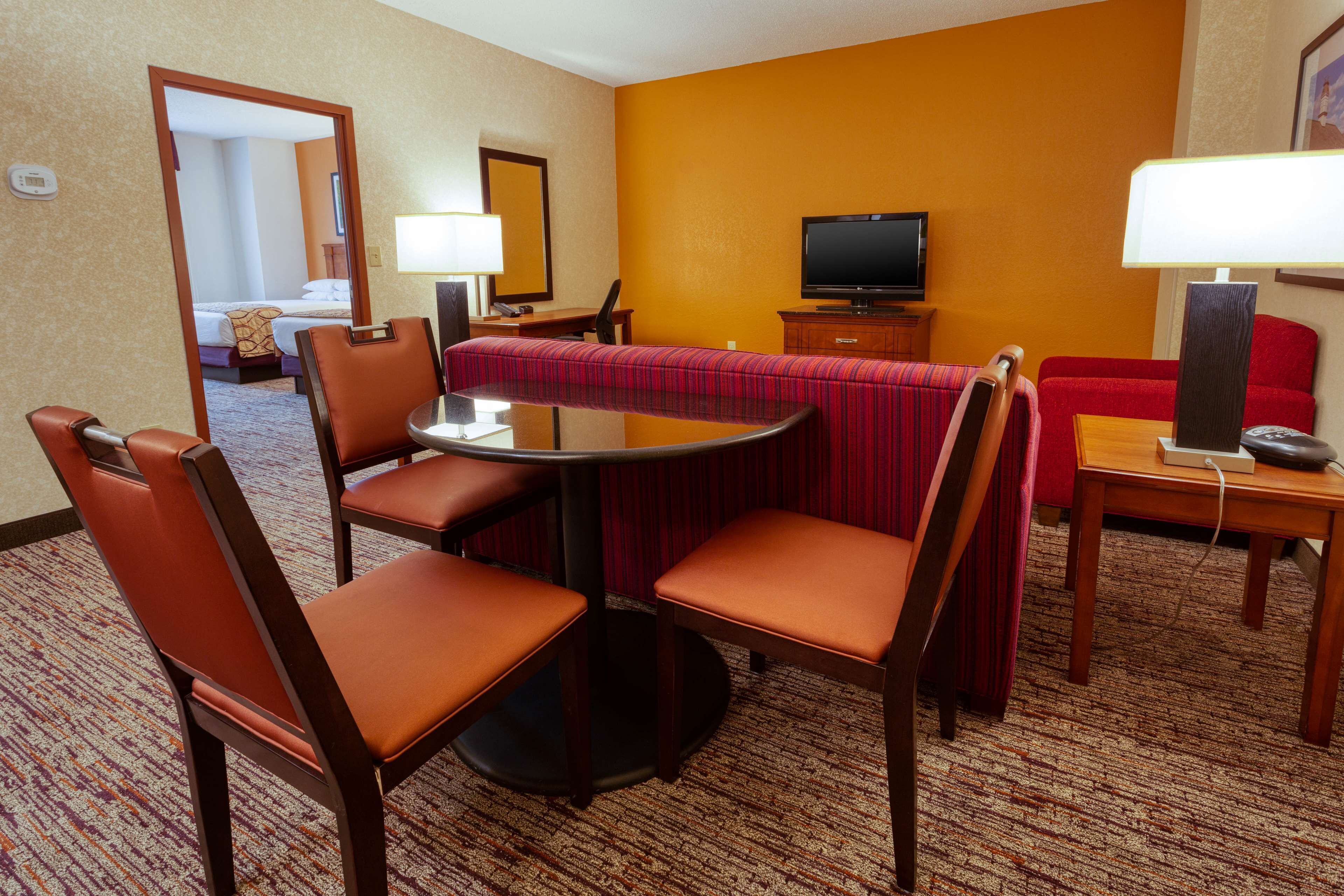 A hotel room with a dining area featuring a round table and four chairs, a television, a red sofa, and a carpeted floor.