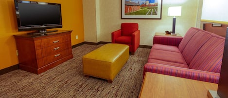 A hotel room with a flat-screen TV, a red armchair, a yellow ottoman, a striped sofa, and a framed picture on the wall.
