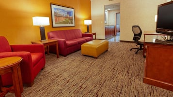 A hotel room with a red sofa, a wooden coffee table, a TV, and a desk with a lamp.