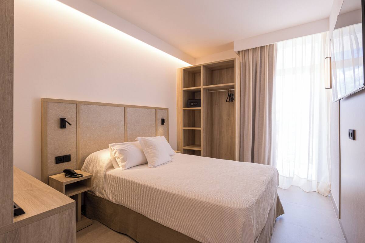 A hotel room with a bed, bedside table, wardrobe, and a large window with curtains.