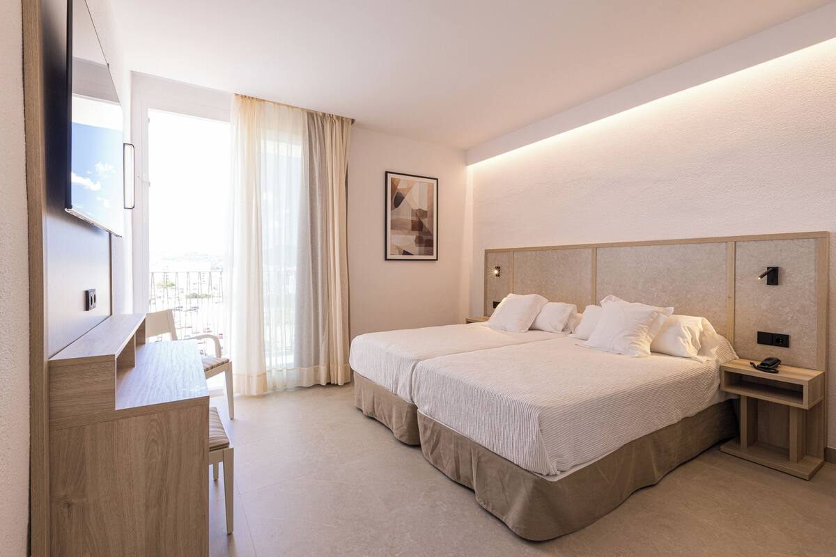 A hotel room with a bed, a desk, a chair, a TV, a bedside table, and a window with curtains.