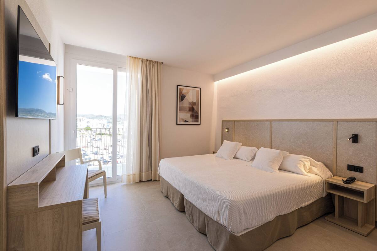 A hotel room with a bed, a desk, a chair, a TV, a bedside table, and a window with curtains.