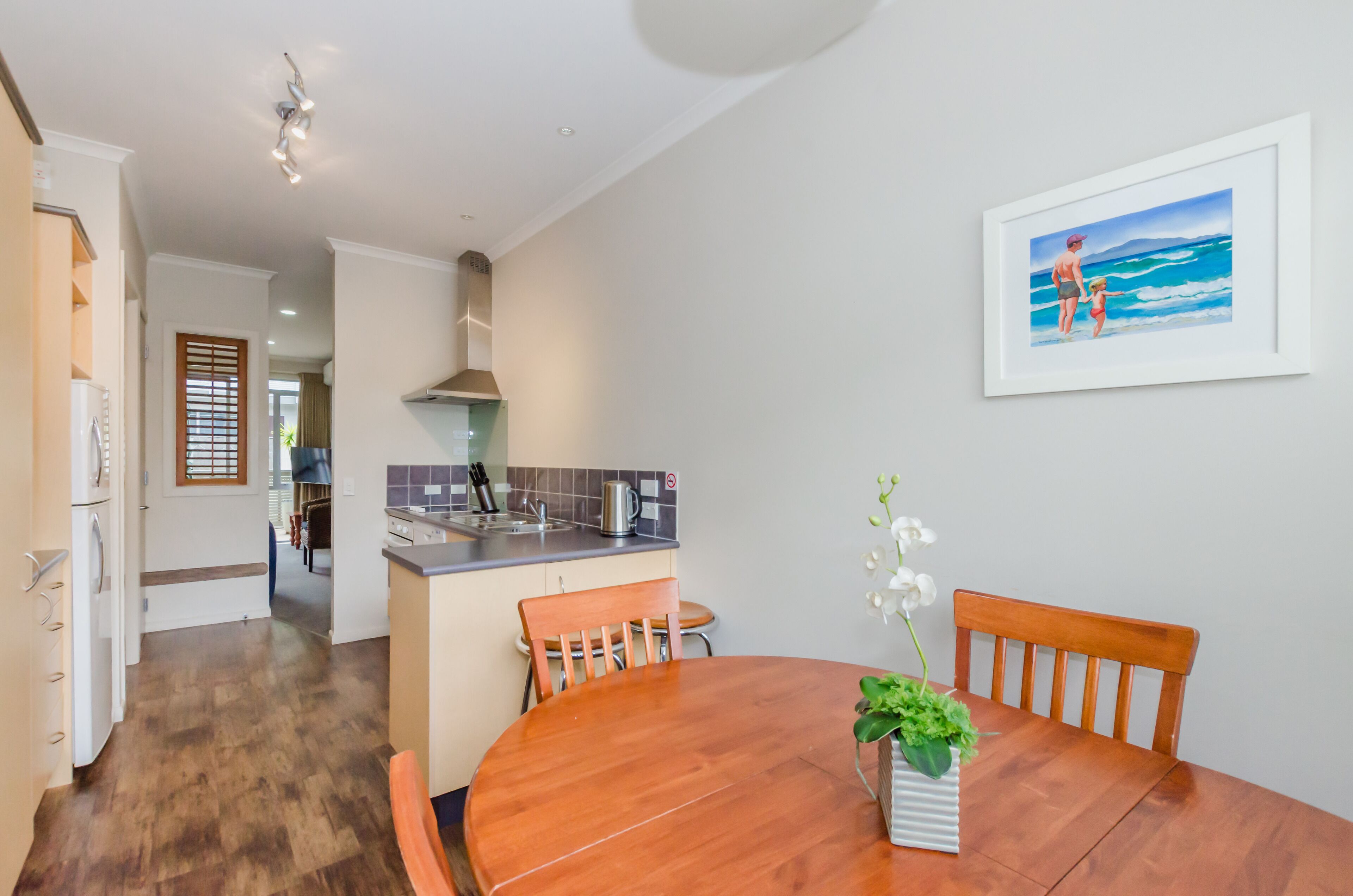 A modern kitchen with a central island, wooden dining table, and chairs. There is a framed picture on the wall and a potted plant on the table.