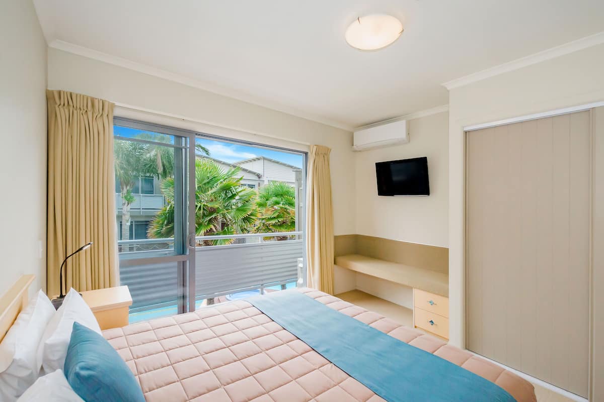 Una habitación de hotel con cama, televisión, puerta corrediza a un balcón y ventana con cortinas.