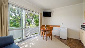 Habitación compacta con mesa de comedor y sillas, un refrigerador pequeño, un televisor de pantalla plana en la pared y un ventanal corredero que da a un balcón con vistas a árboles.
