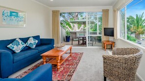 A living room with a blue sofa, a wooden coffee table, a wicker chair, a TV, and a large window with a view of palm trees. - Raumati Sands Resort (Paraparaumu)