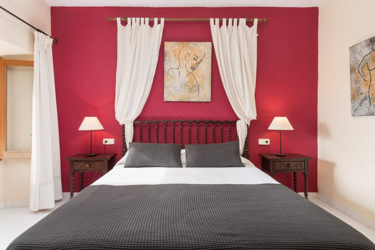 A bedroom with a large bed, two bedside tables with lamps, a red accent wall, and artwork above the bed.