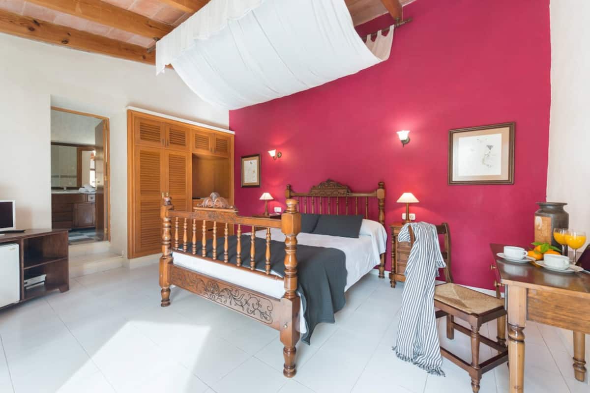 A bedroom with a wooden bed, a red wall, a wooden wardrobe, a desk with a chair, and a bedside table with a lamp.
