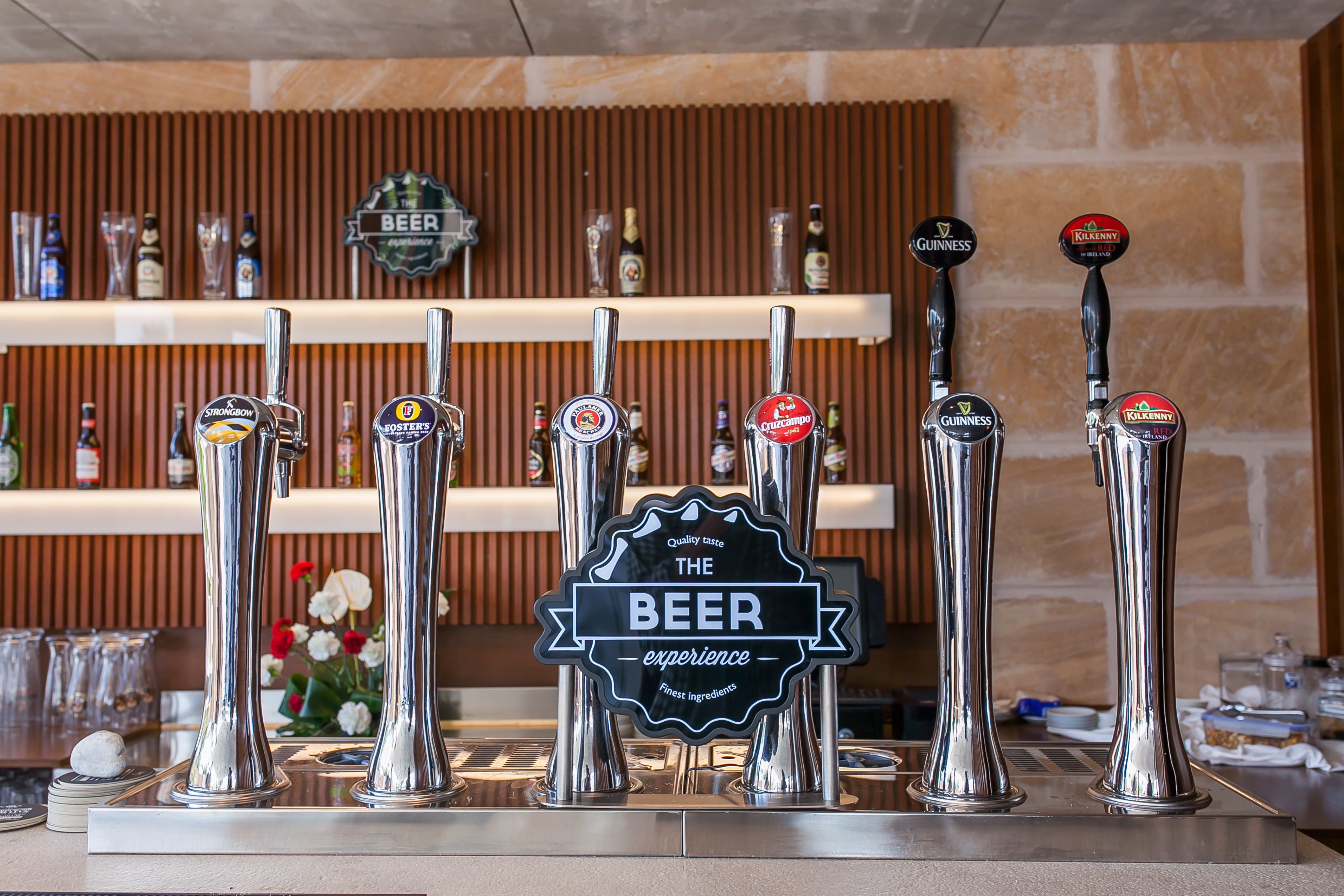 Un bar doté de nombreux robinets à bière, une enseigne indiquant « The Beer Experience », et des étagères remplies de bouteilles en arrière-plan.