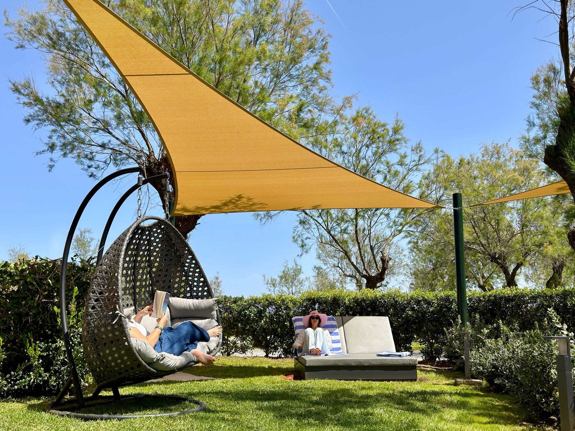 Un jardin avec une chaise suspendue et un fauteuil de relaxation, le tout installé sous une voile d’ombrage jaune.