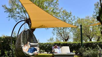 Un jardin avec une chaise suspendue et un fauteuil de relaxation, le tout installé sous une voile d’ombrage jaune.