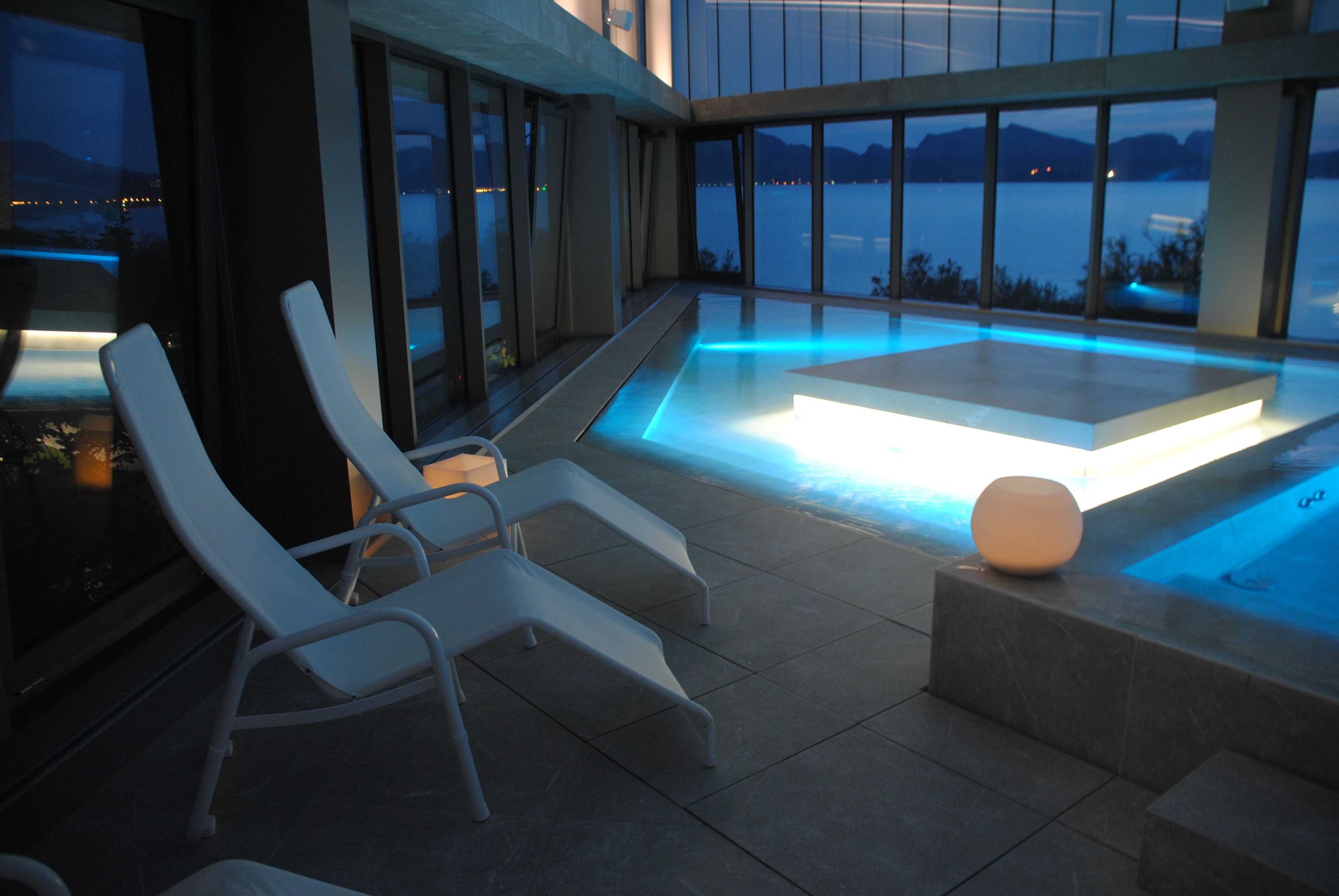 A modern outdoor pool area with lounge chairs and a hot tub, overlooking a body of water.
