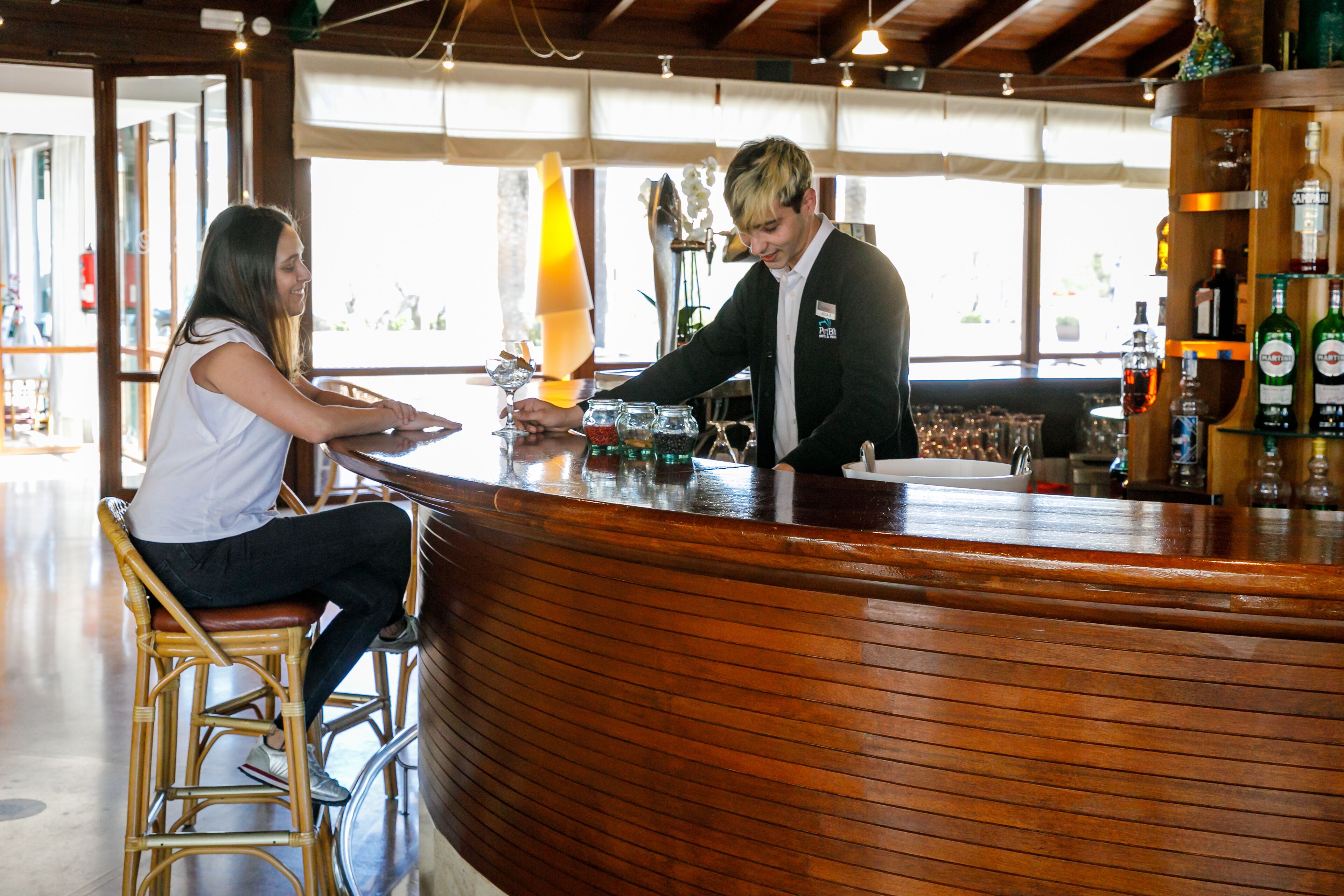 Eine Bar mit einer gebogenen Holztheke, einer Frau, die auf einem Hocker sitzt, und einem Barkeeper, der Getränke zubereitet.
