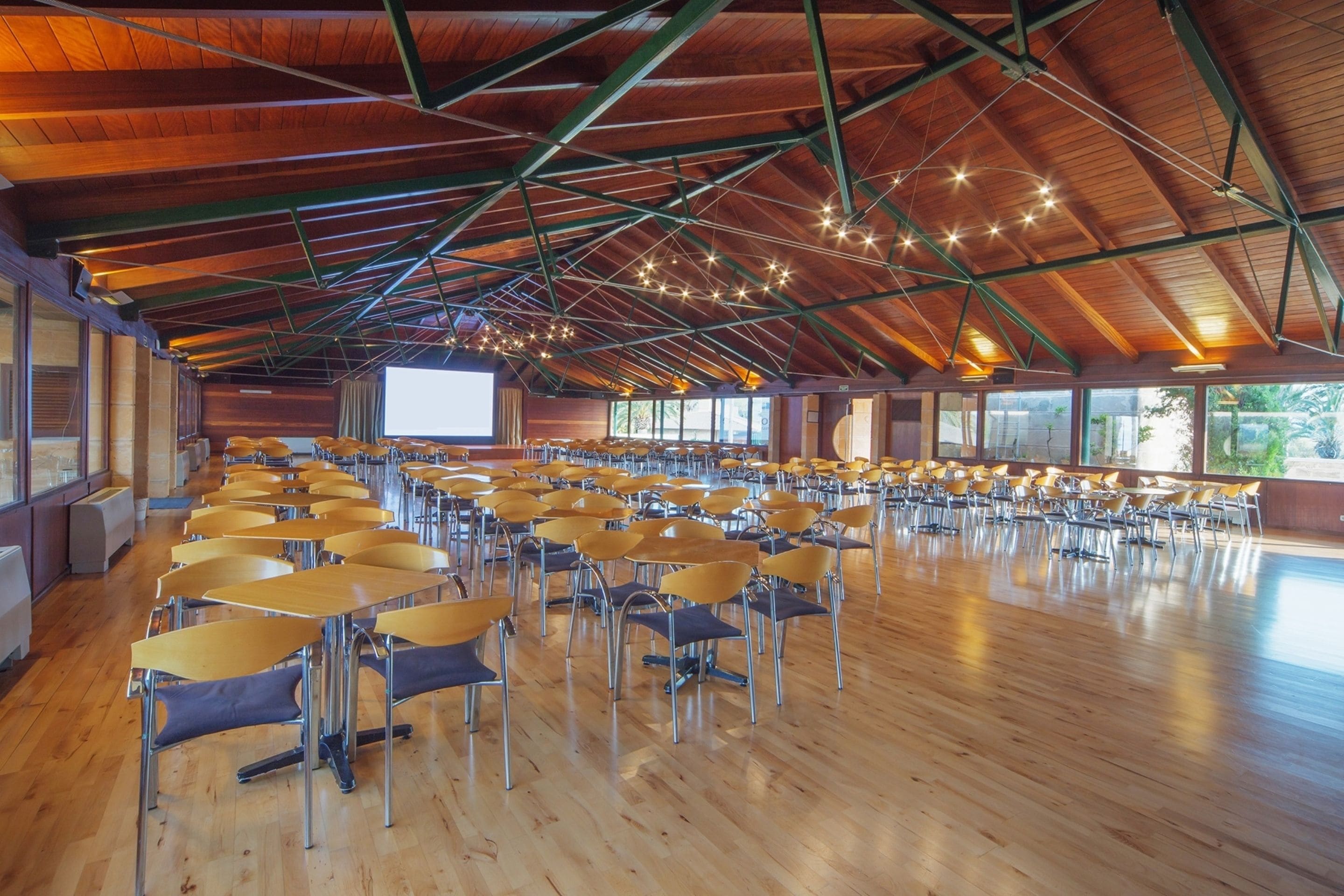 A large hall with rows of tables and chairs, wooden floors, and a high ceiling with exposed beams and hanging lights.