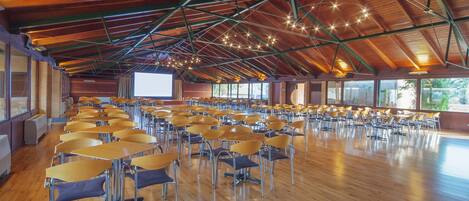 A large hall with rows of tables and chairs, wooden floors, and a high ceiling with exposed beams and hanging lights.