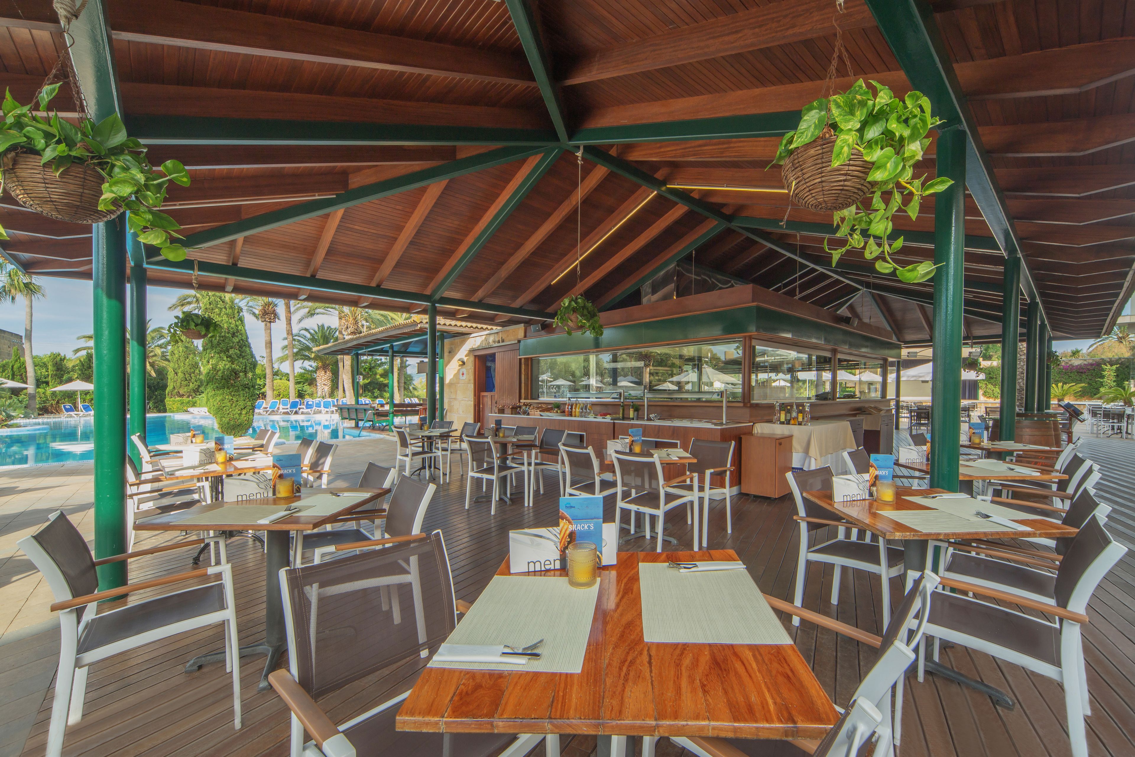 Un restaurante al aire libre con mesas y sillas de madera, plantas colgantes y una piscina al fondo.