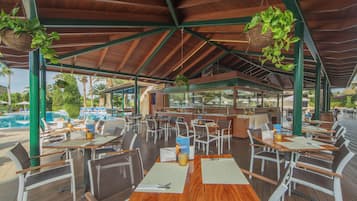 Un restaurante al aire libre con mesas y sillas de madera, plantas colgantes y una piscina al fondo.