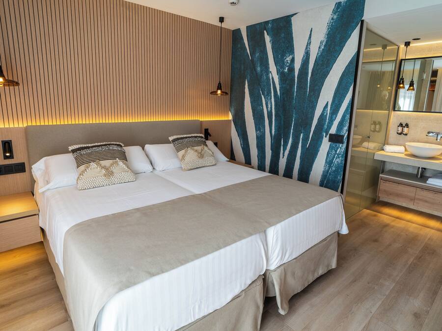 Una habitación de hotel moderna con una cama grande, cabecera de madera, lámpara de pared y baño con lavabo y espejo.