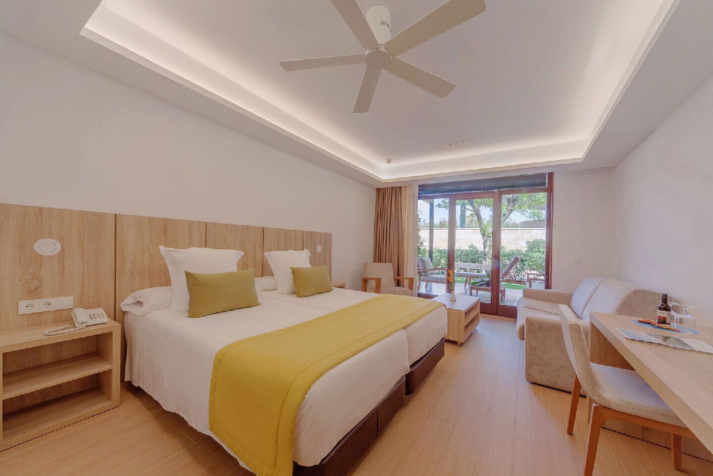 Une chambre d’hôtel moderne, dotée d’un grand lit, d’un bureau et d’une chaise. On y trouve un ventilateur de plafond, une fenêtre avec des rideaux et une vue sur la verdure environnante.