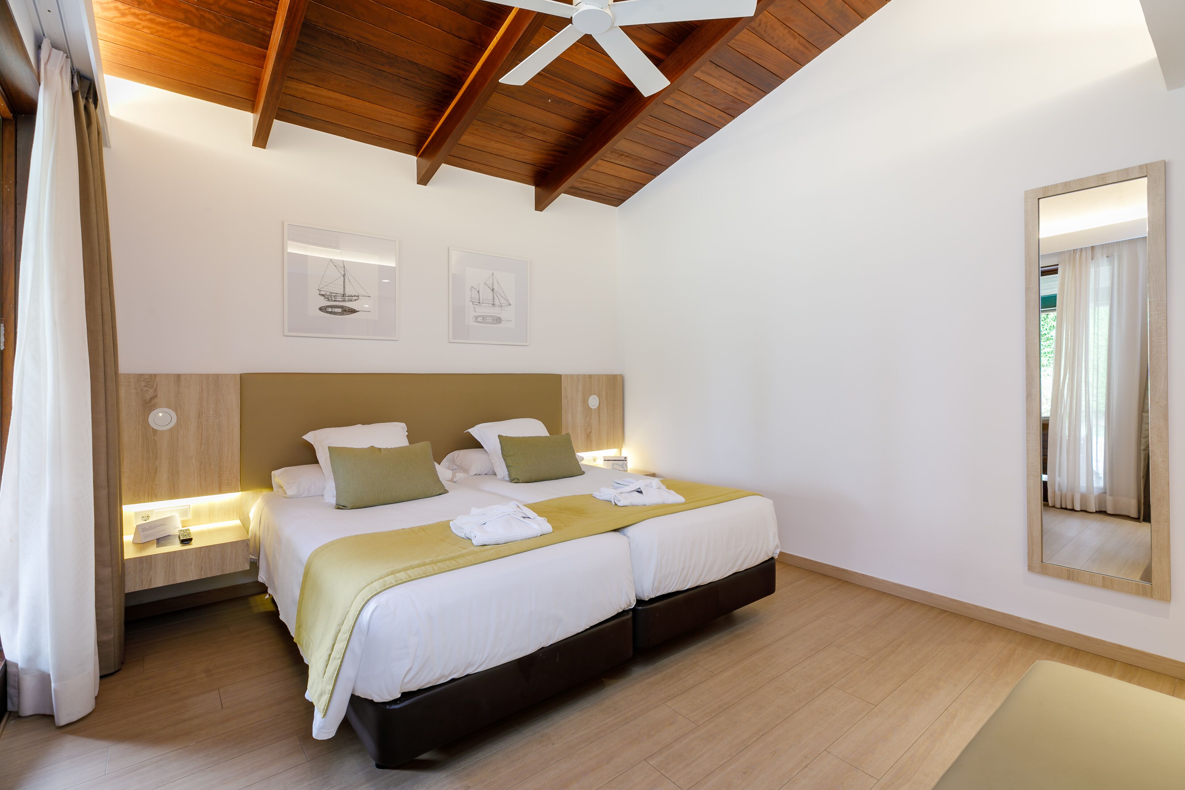 A modern hotel room with a wooden ceiling, a large bed with white bedding and green pillows, a bedside table with a lamp, and a large mirror on the wall.