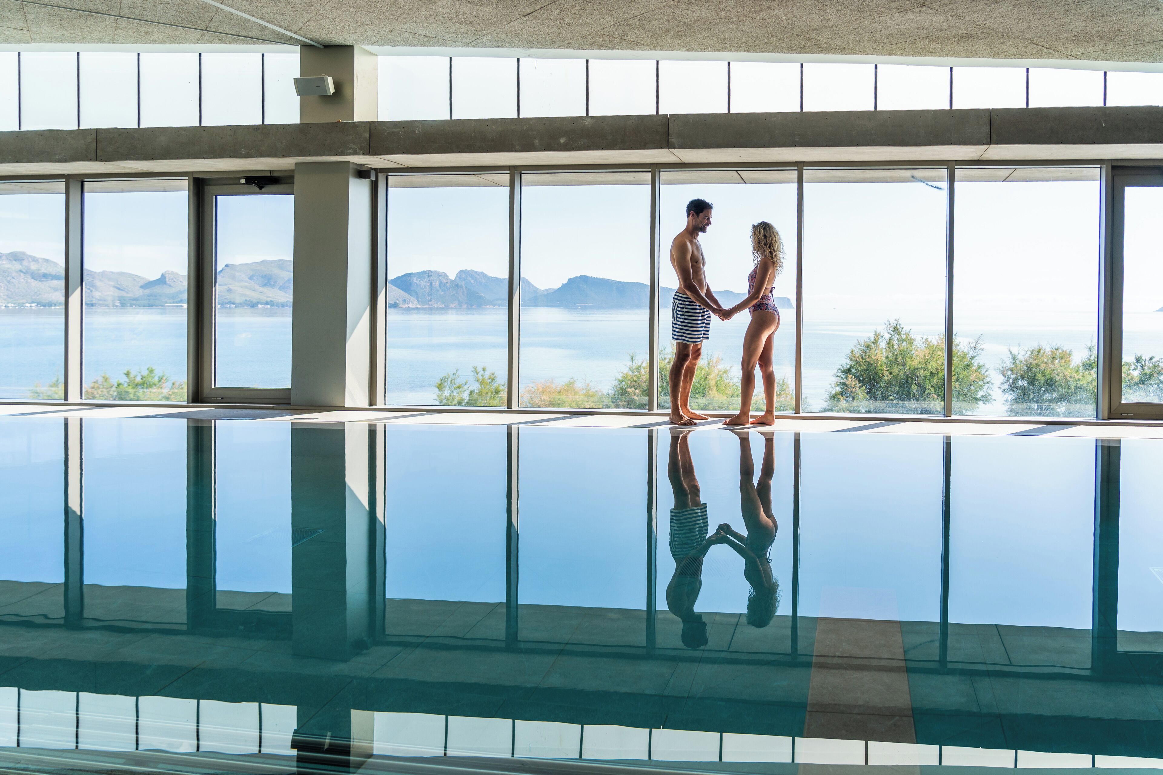 Un couple se tient près d’une grande piscine intérieure, avec vue sur les montagnes et l’eau.