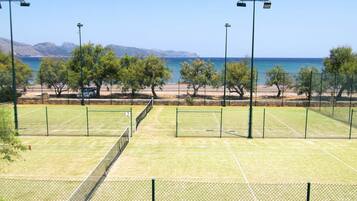 An outdoor tennis court with a net, surrounded by trees and a fence, with a view of the sea and mountains in the background.