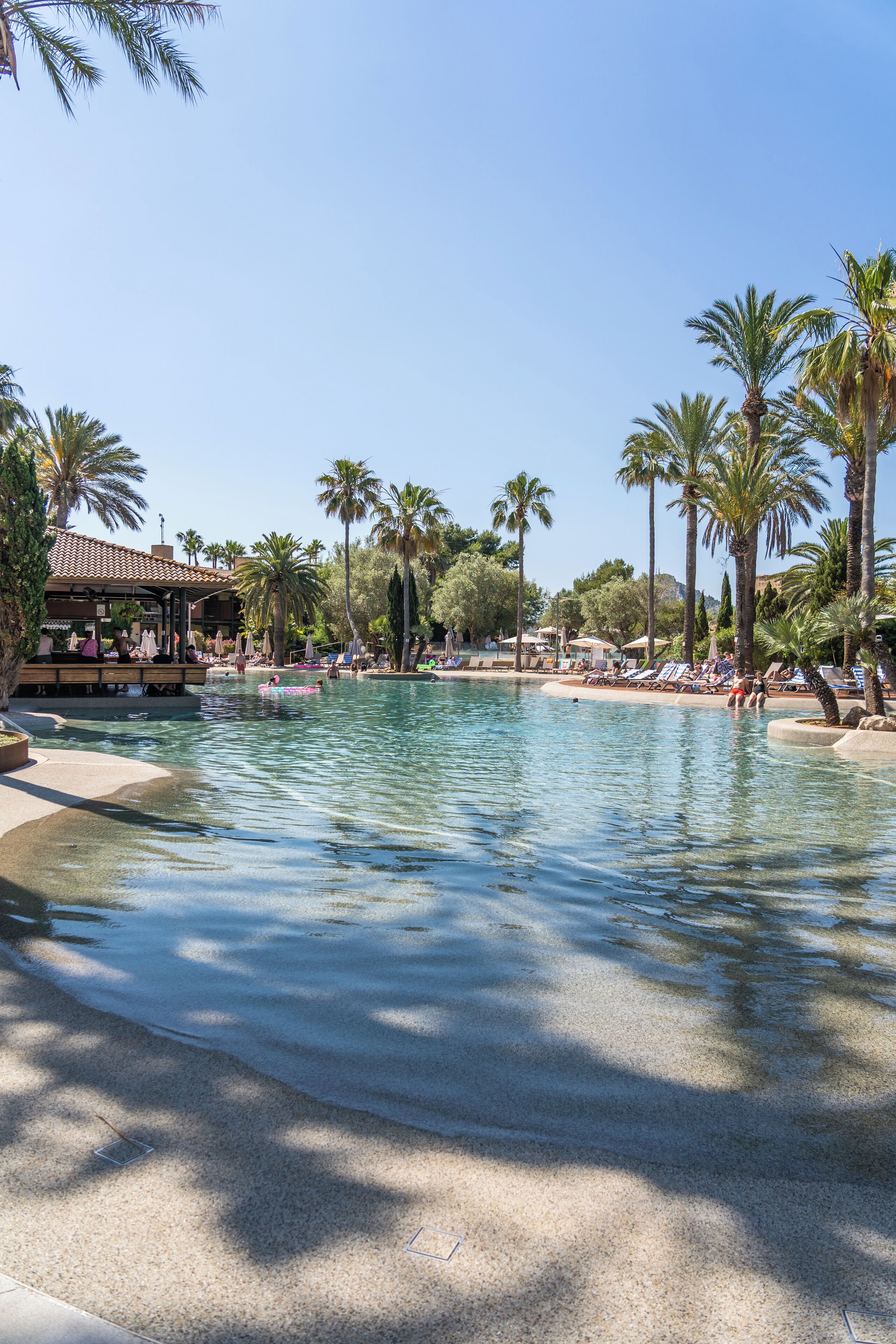 Un espace piscine aménagé dans un complexe hôtelier, avec des palmiers, des chaises longues et un kiosque.