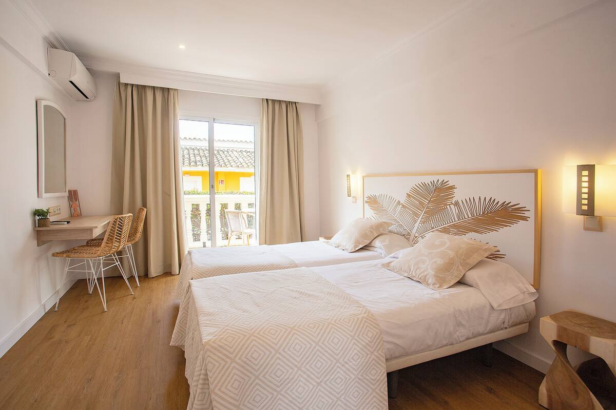 A hotel room with two beds, a desk, a chair, and a view of a yellow building through the window.