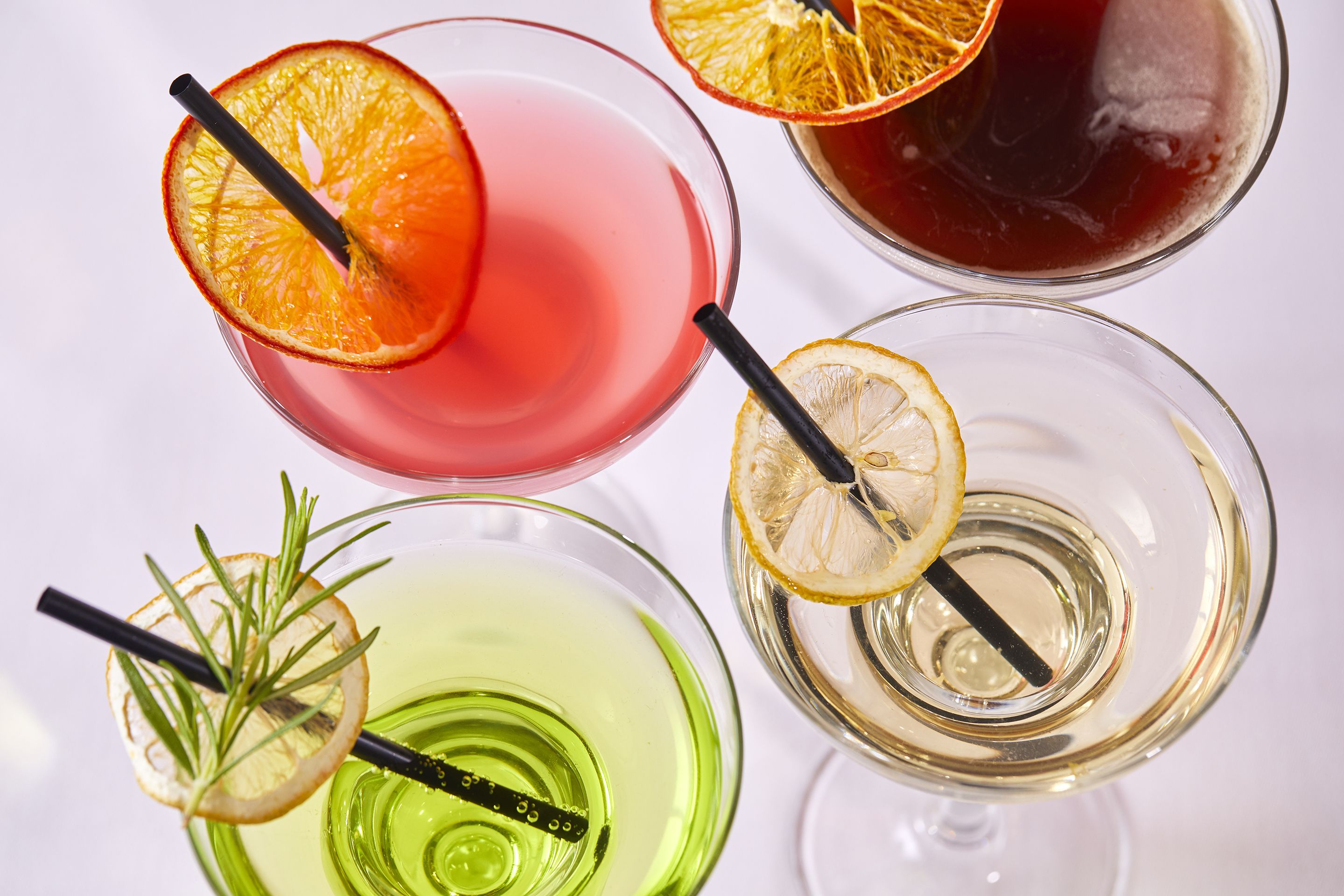 Four glasses of different colored drinks, each with a slice of fruit and a straw.