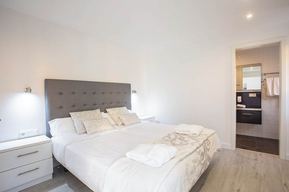 A neatly made bed with a grey headboard, white bedding, and a folded towel. There is a bedside table with drawers and a lamp.