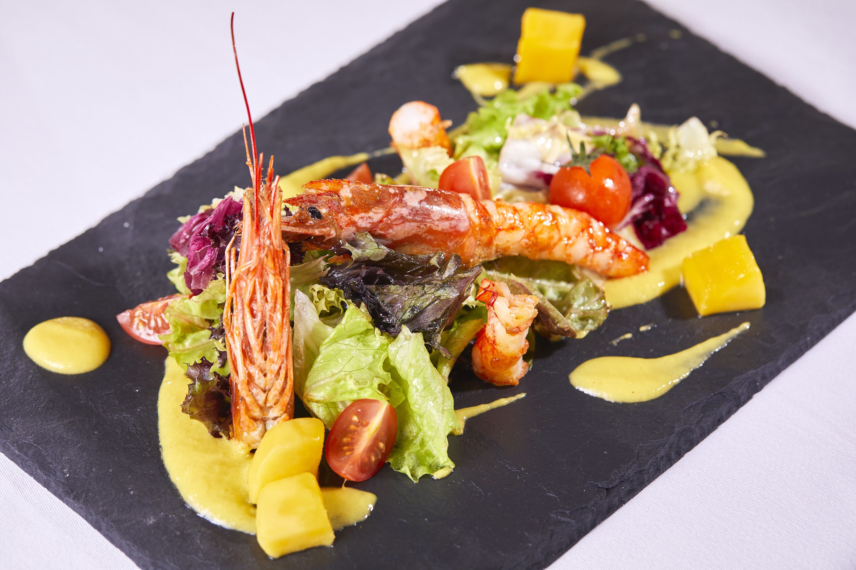A seafood dish with shrimp, vegetables, and mango on a black slate plate.