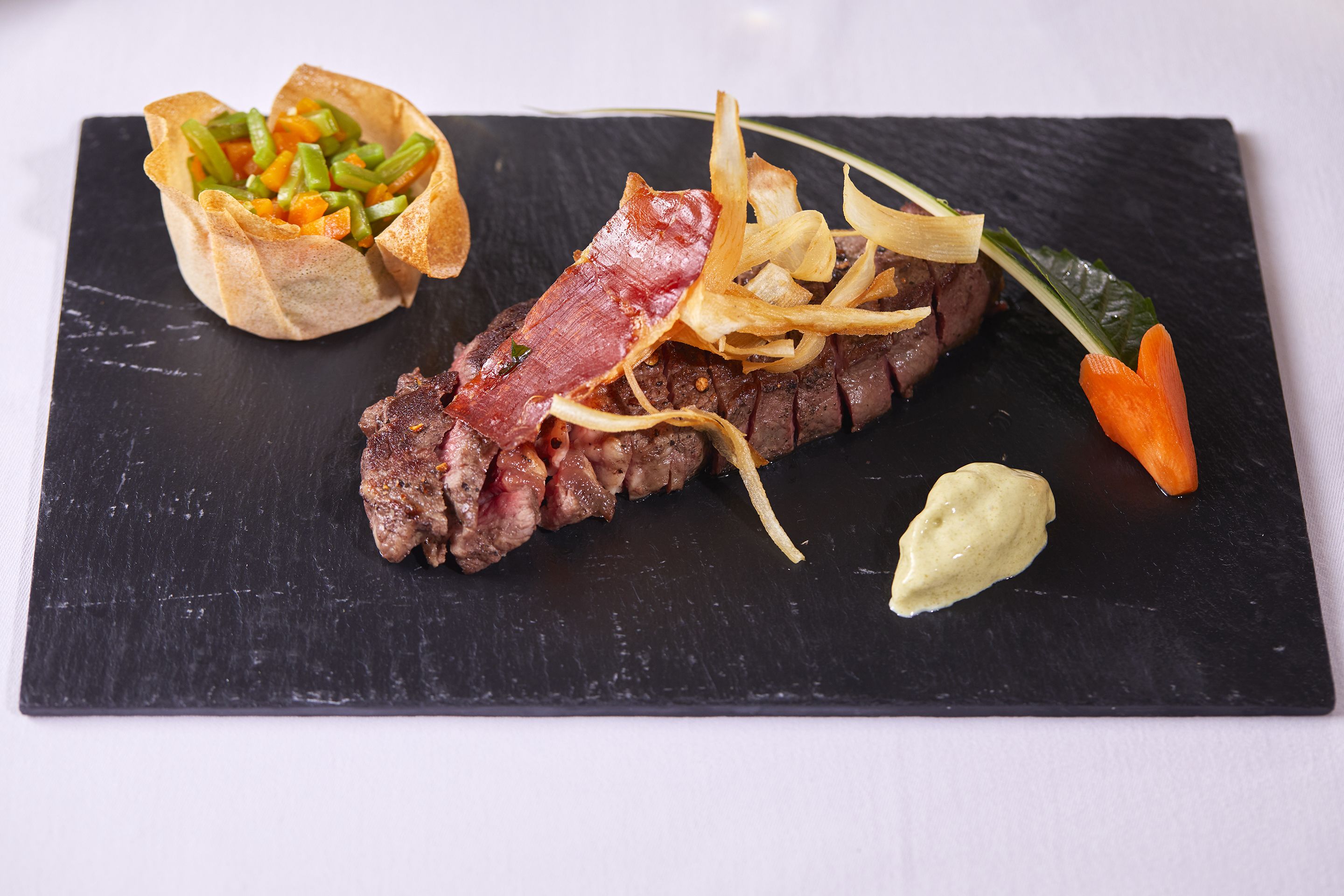 Une planche de service en ardoise noire sur laquelle est présentée une portion de steak garnie de lardons, un petit bol de légumes et une cuillerée de sauce.