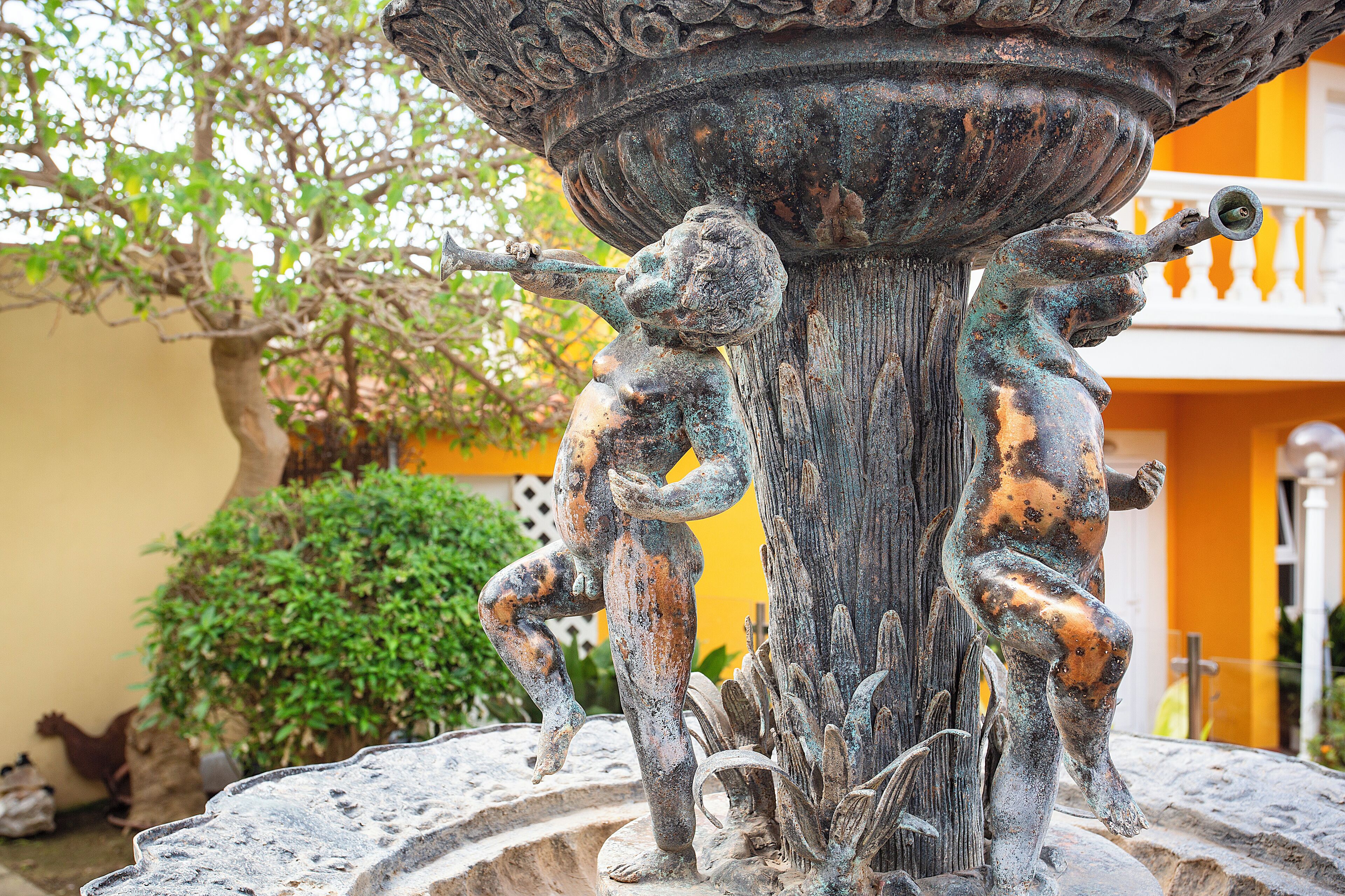 Una fuente de bronce con dos figuras, rodeada de follaje y un edificio amarillo al fondo.