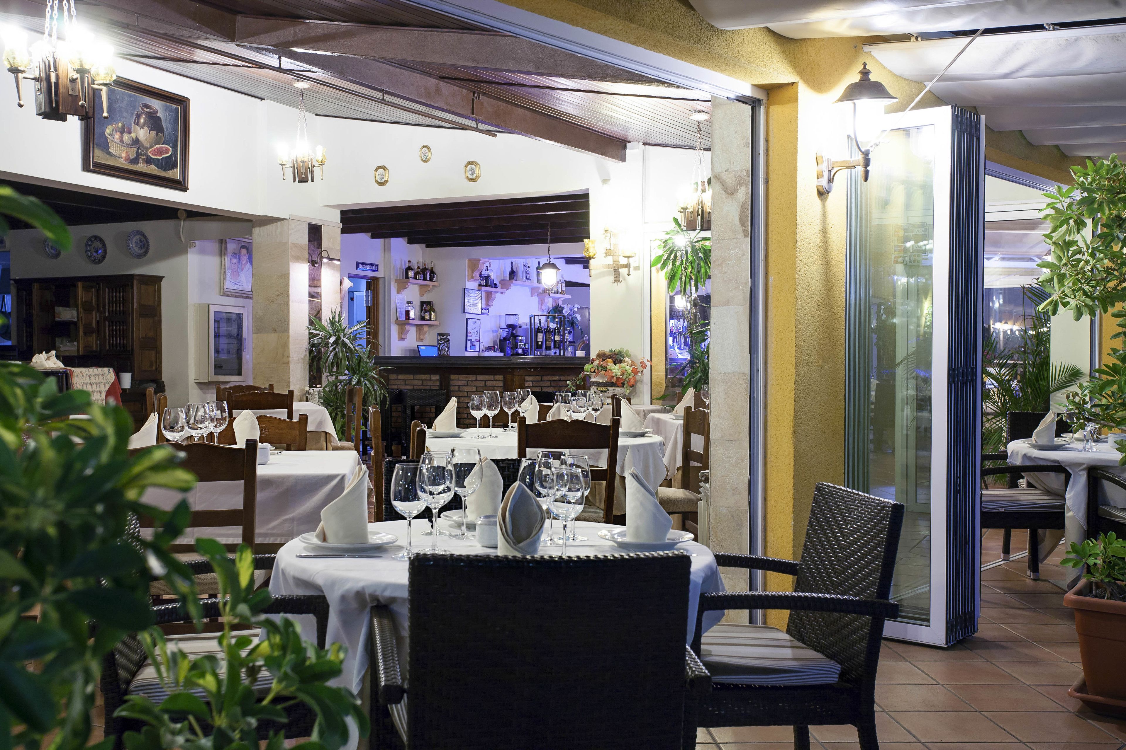 Un comedor interior con mesas dispuestas para dos, sillas y una barra visible al fondo.