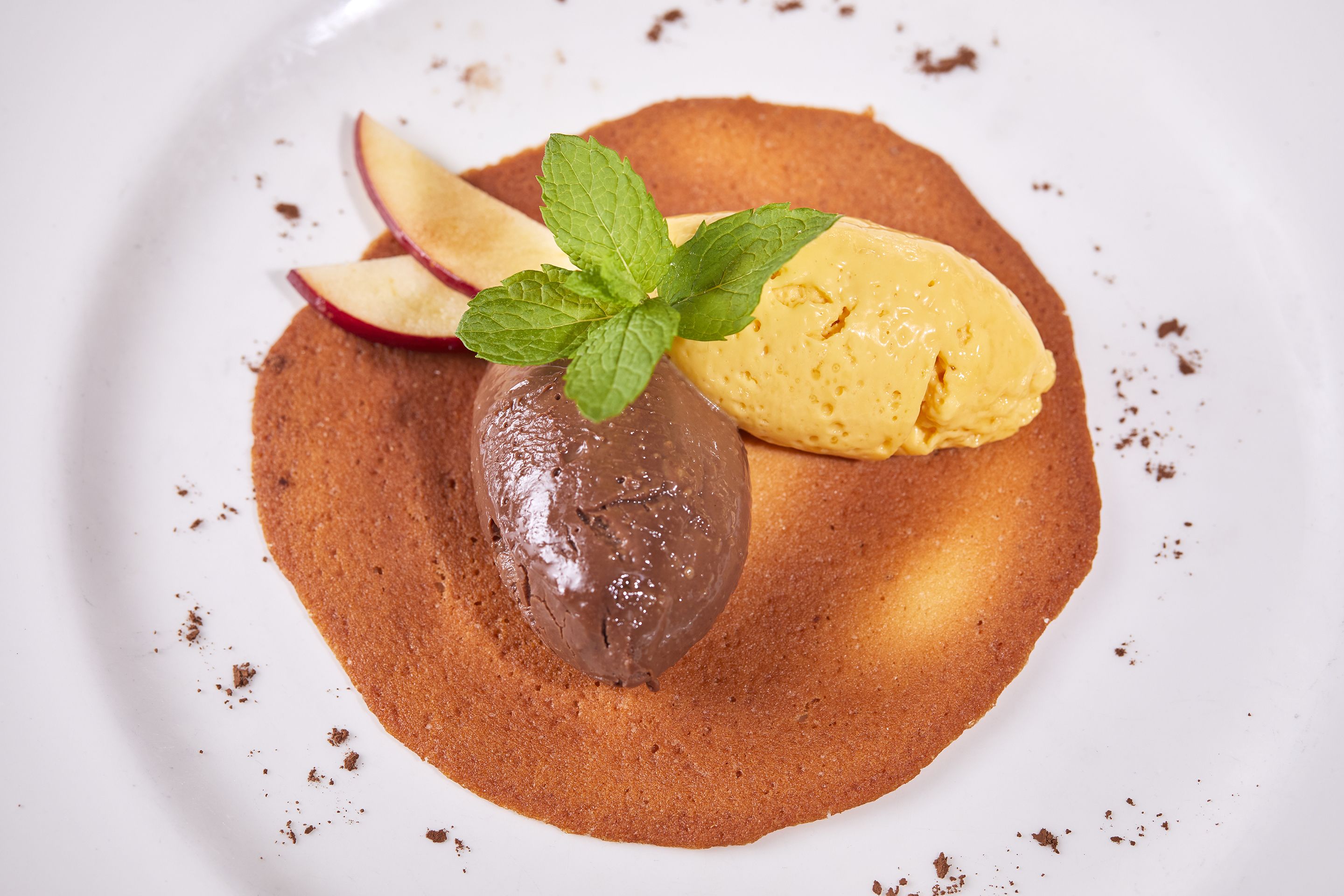 Un postre con helado de chocolate y vainilla, rodajas de manzana y hojas de menta sobre un plato blanco.