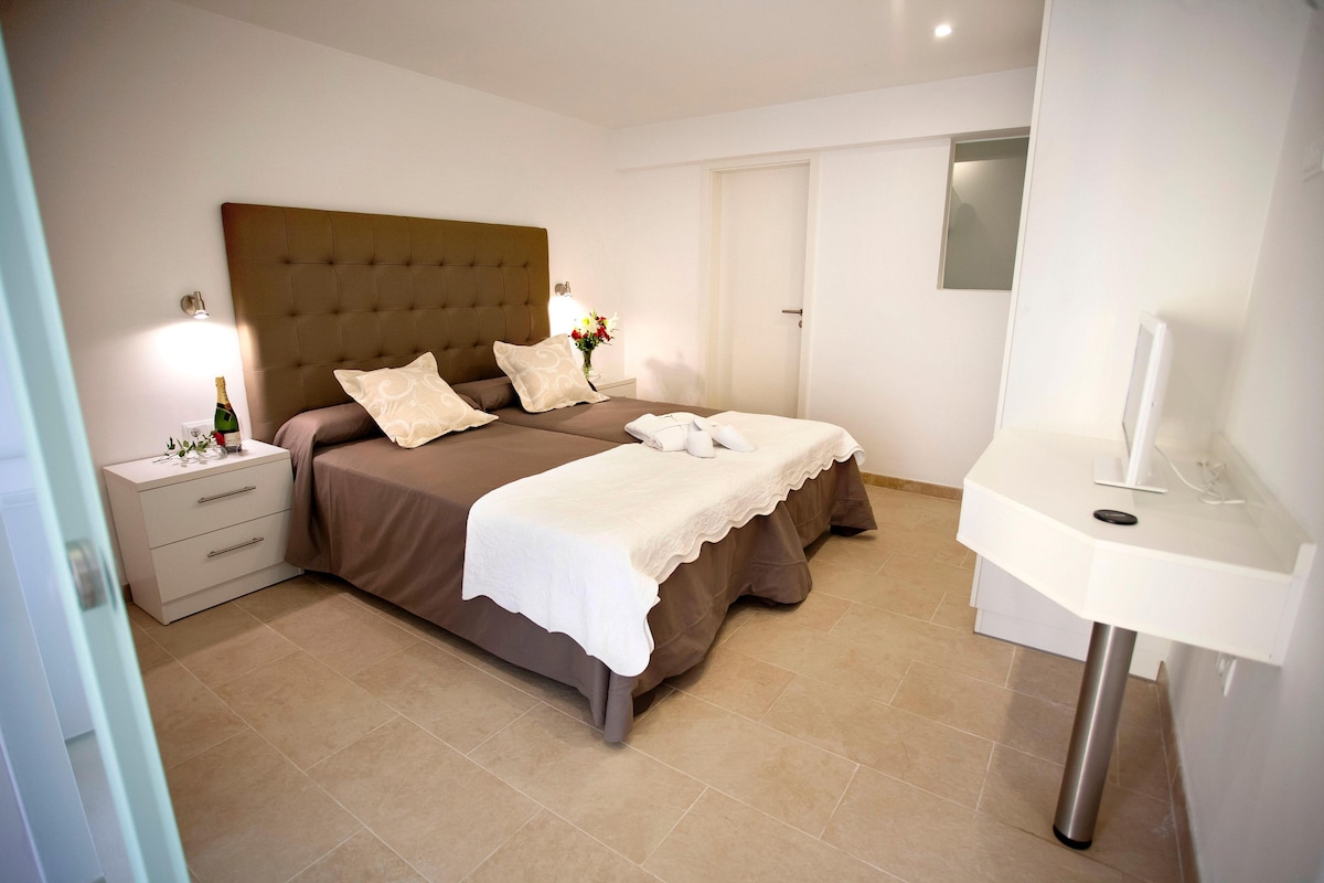 A hotel room with a double bed, a bedside table with a bottle and glasses, a small nightstand, a white bathroom with a sink and mirror.