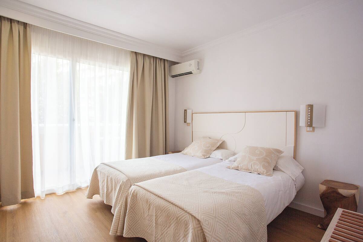 A hotel room with two beds, a wooden floor, a window with sheer curtains, an air conditioning unit, and a bedside table.