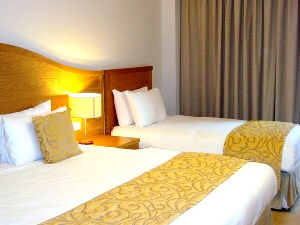 A hotel room with two beds, a wooden headboard, a nightstand with a lamp, and a curtain. - Parkview Hotel (Newtownmountkennedy)