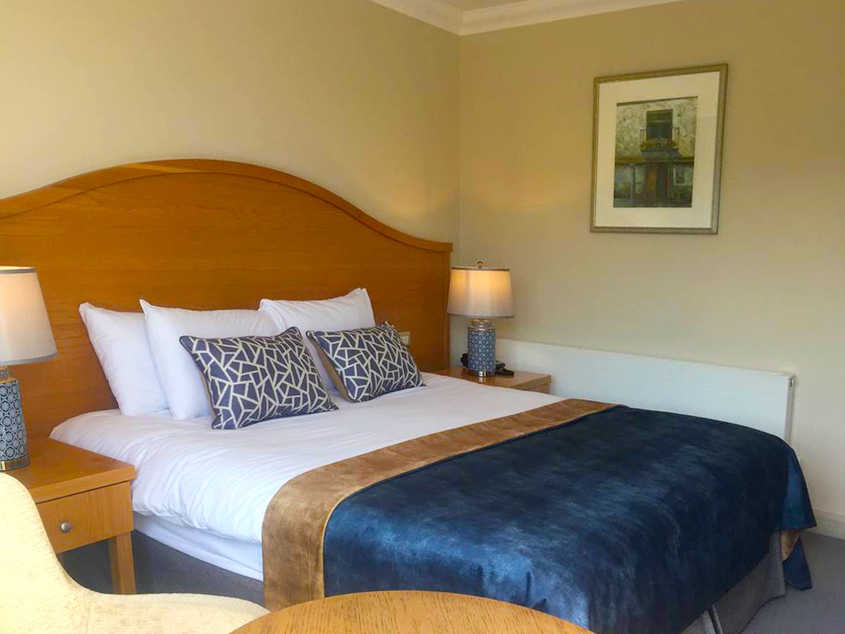 Ein Hotelzimmer mit Holz Kopfteil, einem großen Bett mit weißen und gemusterten Kissen, zwei Nachttischlampen und einem gerahmten Bild an der Wand.