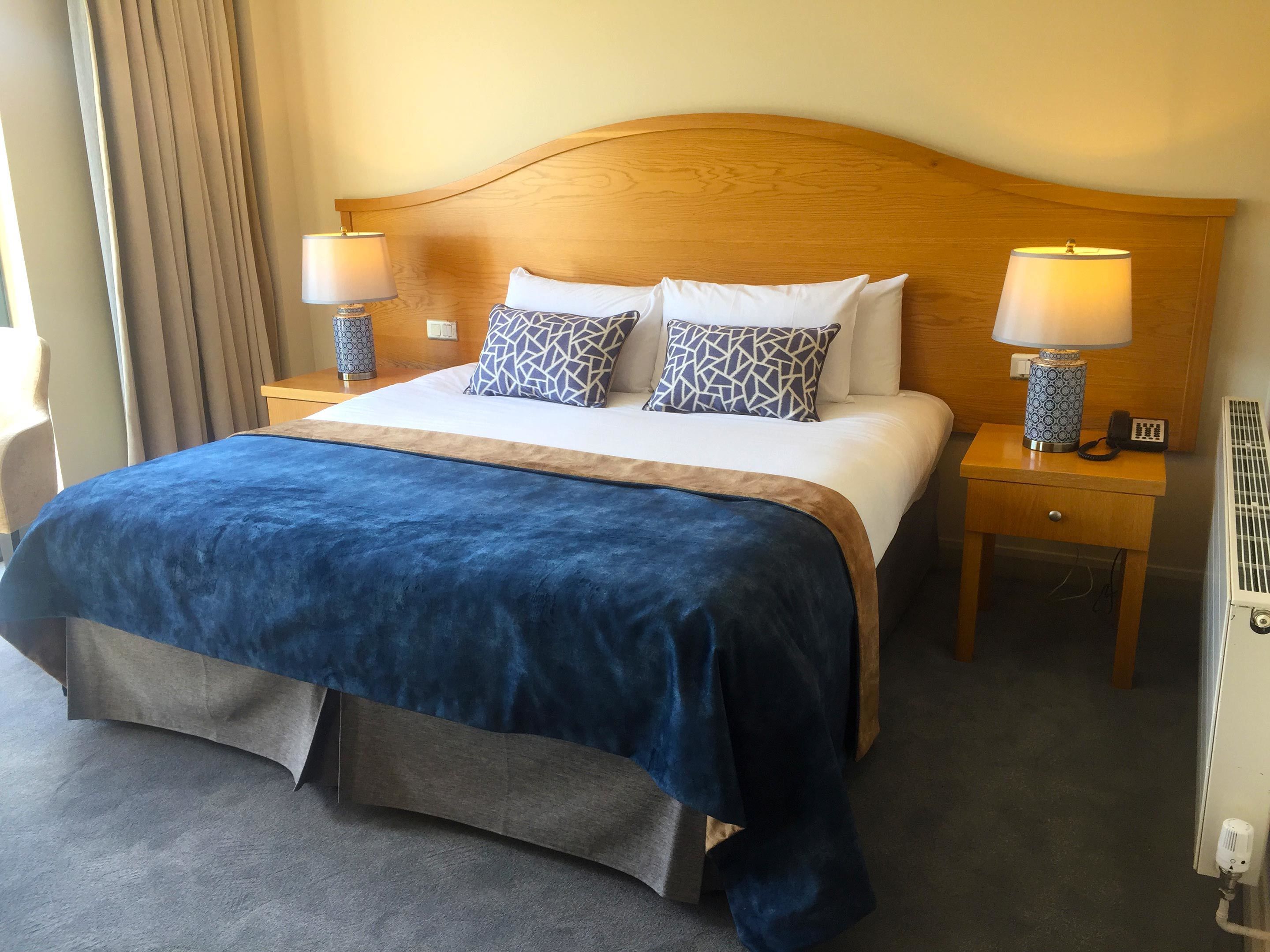 A hotel room with a large bed, two bedside tables with lamps, a wooden headboard, and a grey carpet.