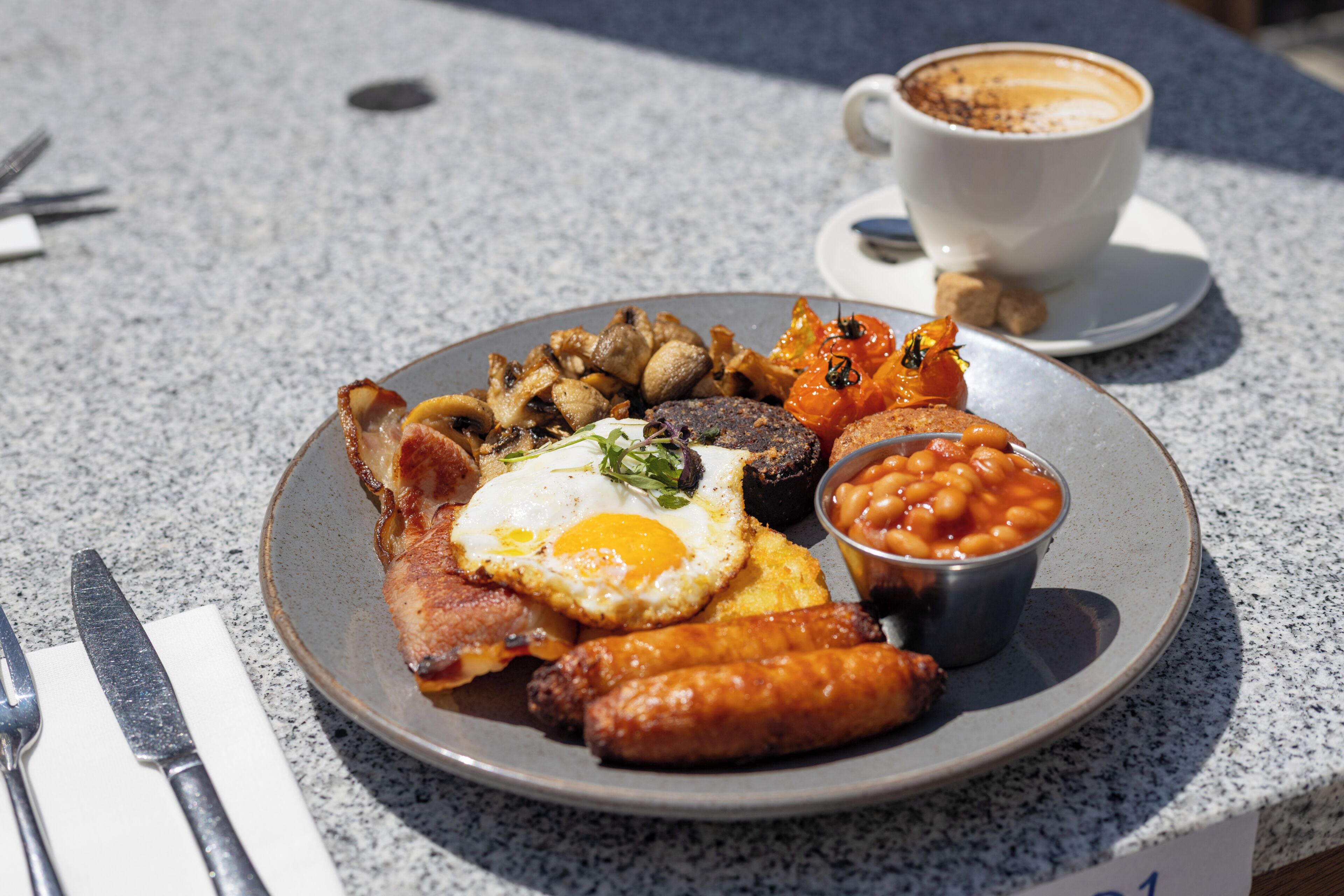 A plate of breakfast food including eggs, sausages, beans, and toast, with a cup of coffee on the side.
