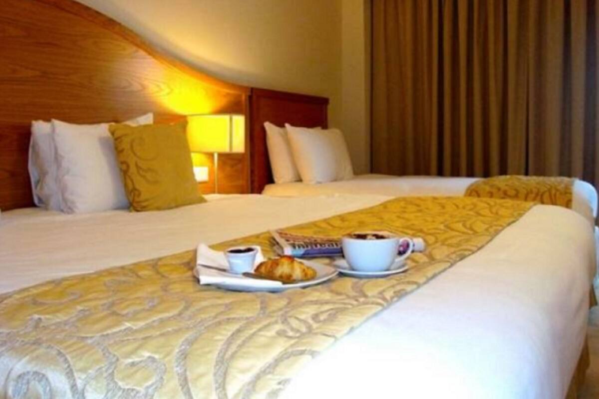Une chambre d’hôtel avec un lit soigneusement fait, une table de chevet avec une lampe et un plateau avec des croissants et du café.