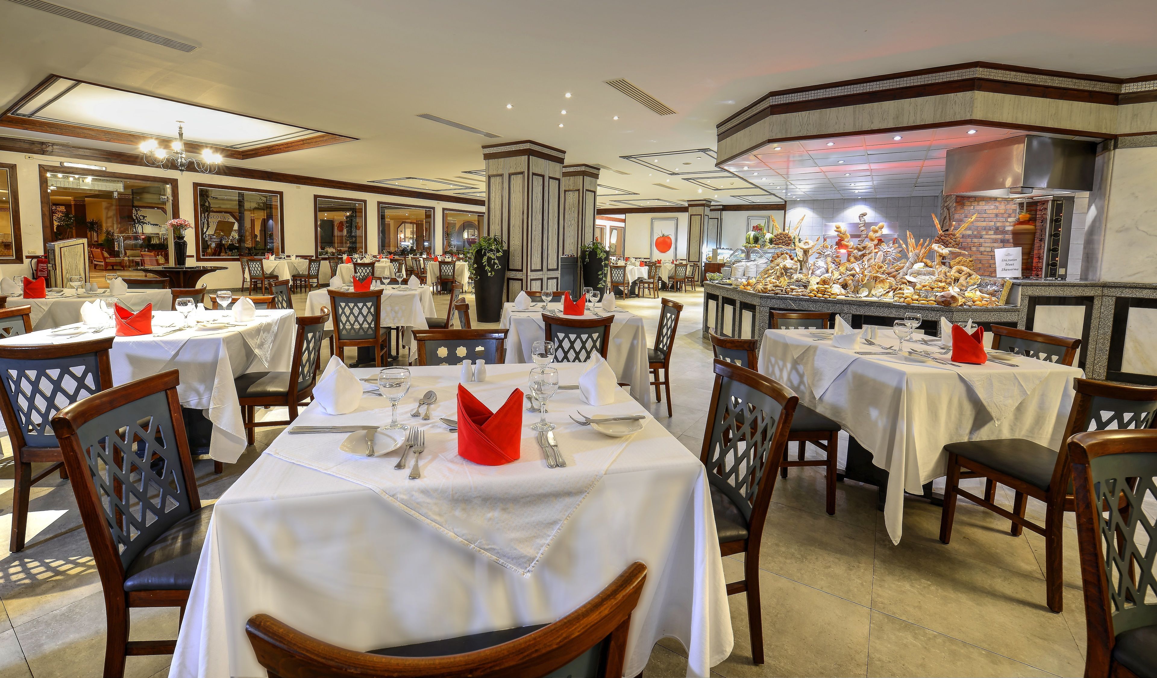 Un restaurant doté de nombreuses tables dressées pour le service, avec une décoration centrale remarquable et une cuisine visible au fond de la salle.