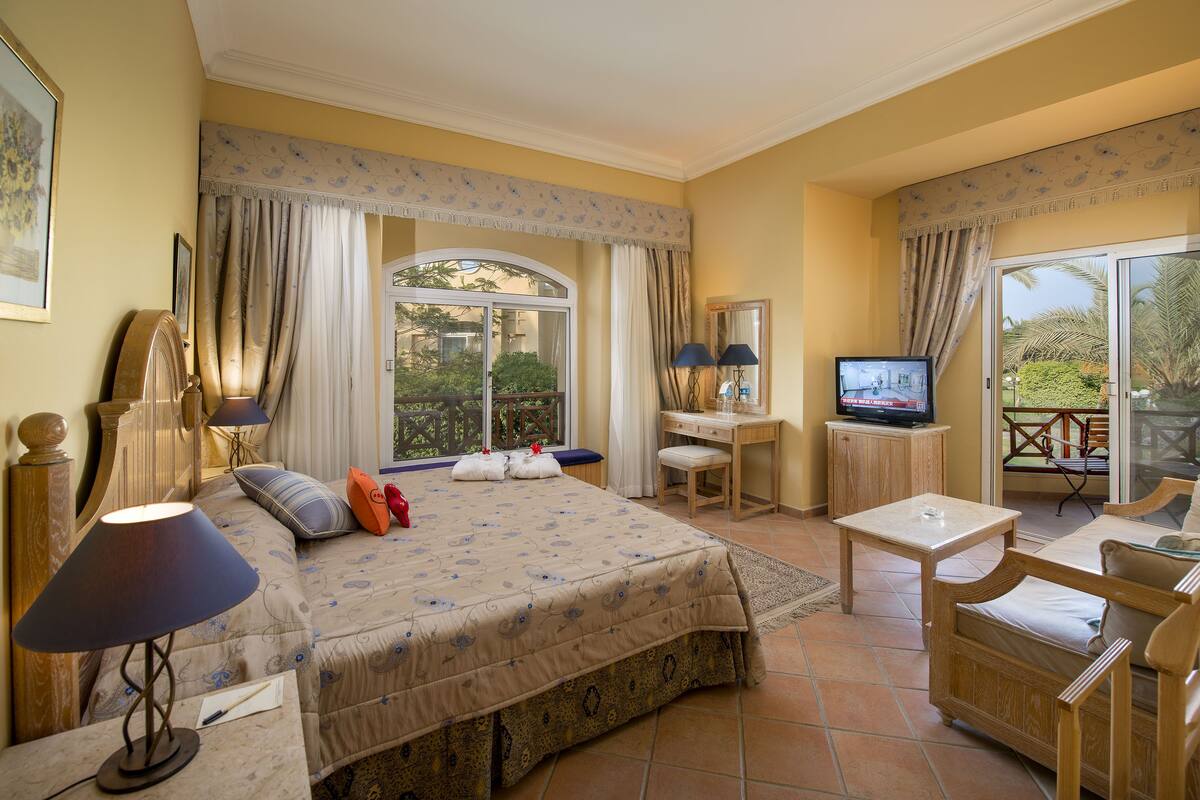 Une chambre à coucher comprenant un lit, des lampes de chevet, un bureau, une télévision et un balcon aménagé avec du mobilier d’extérieur.