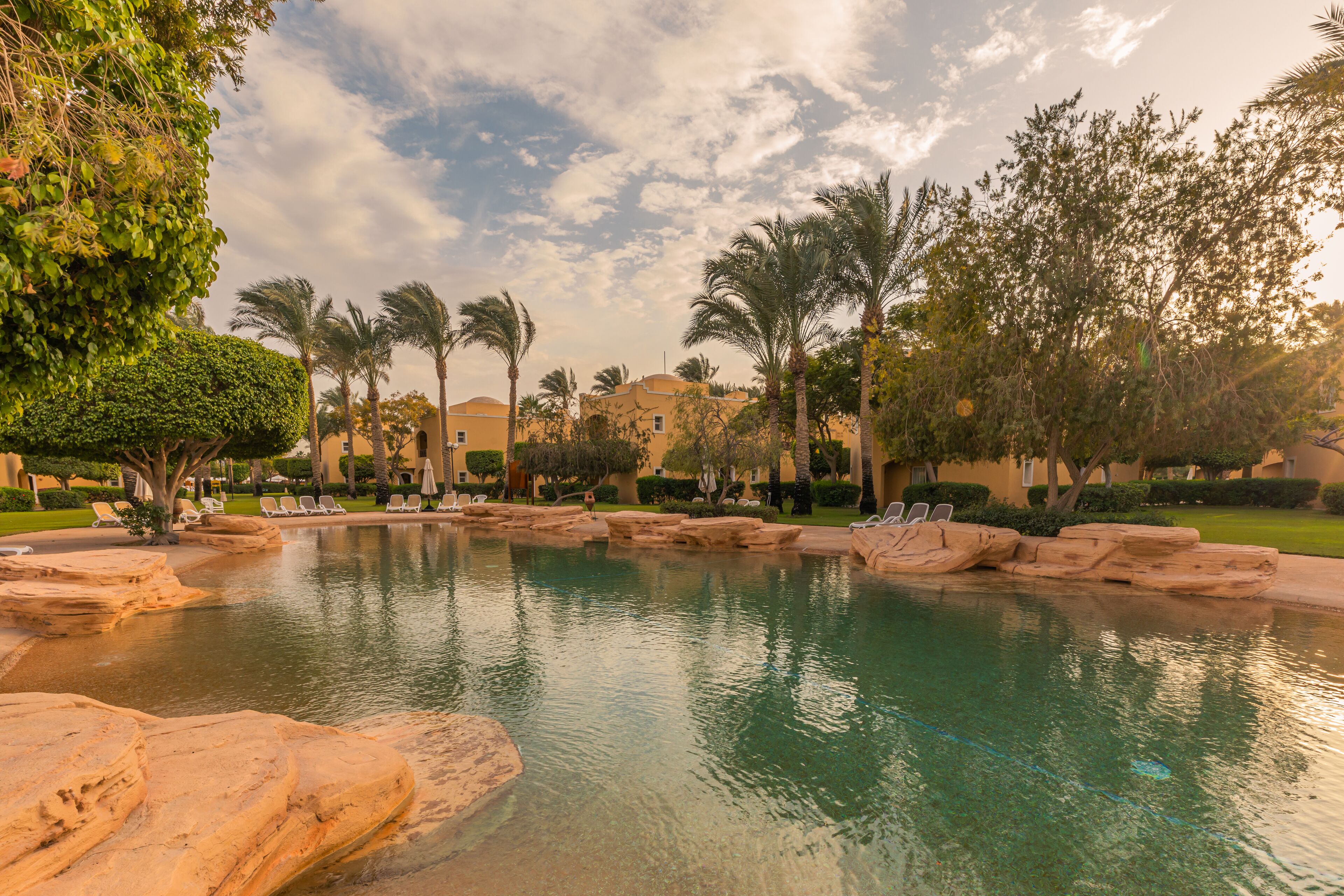 Un área de piscina con agua cristalina, rodeada de rocas y palmeras, con edificios al fondo.