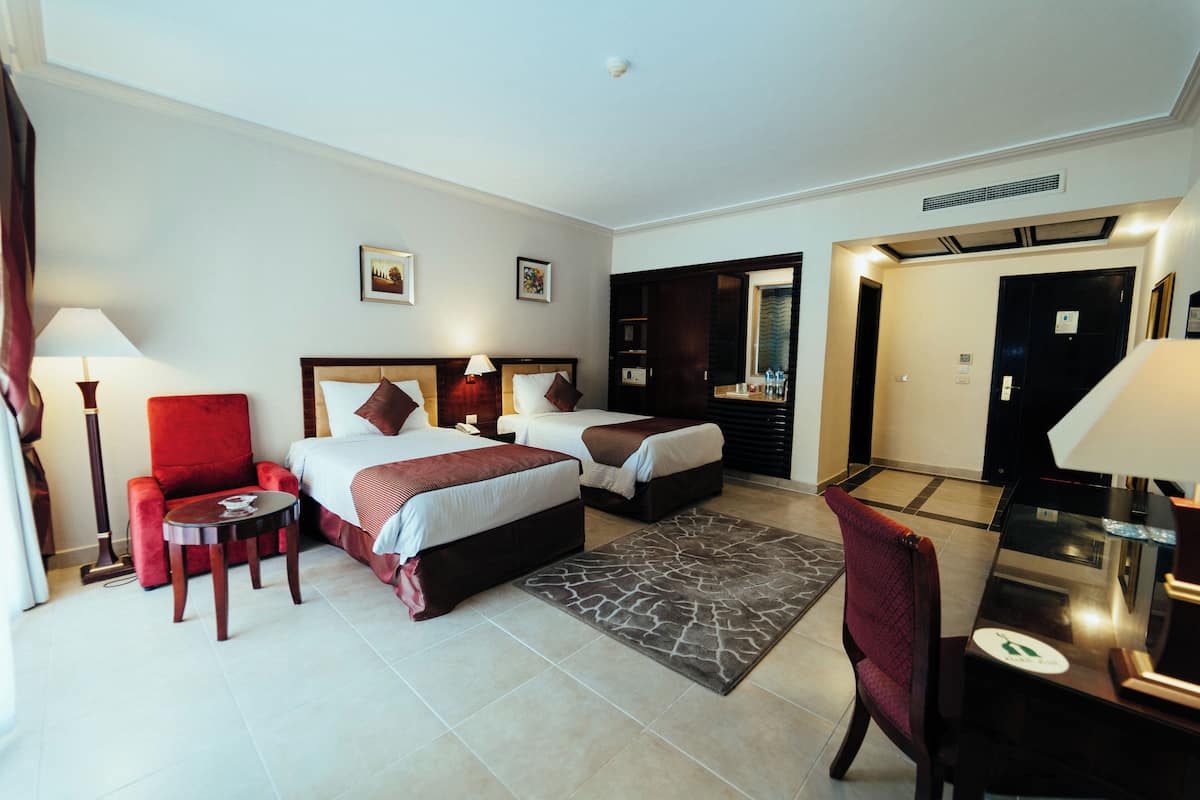 Une chambre d’hôtel avec deux lits, un fauteuil rouge, une petite table ronde et un bureau avec une chaise.