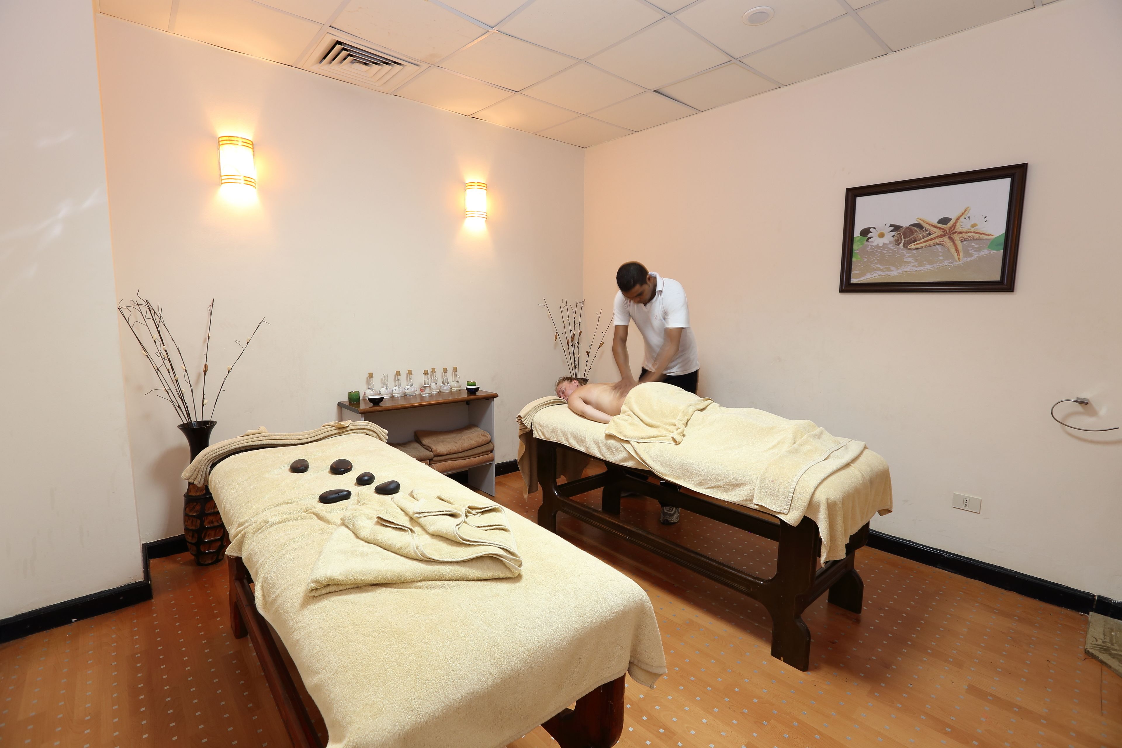 Une salle de spa équipée de deux tables de massage, d’un tableau accroché au mur et d’une étagère où sont posées des bouteilles et un vase.