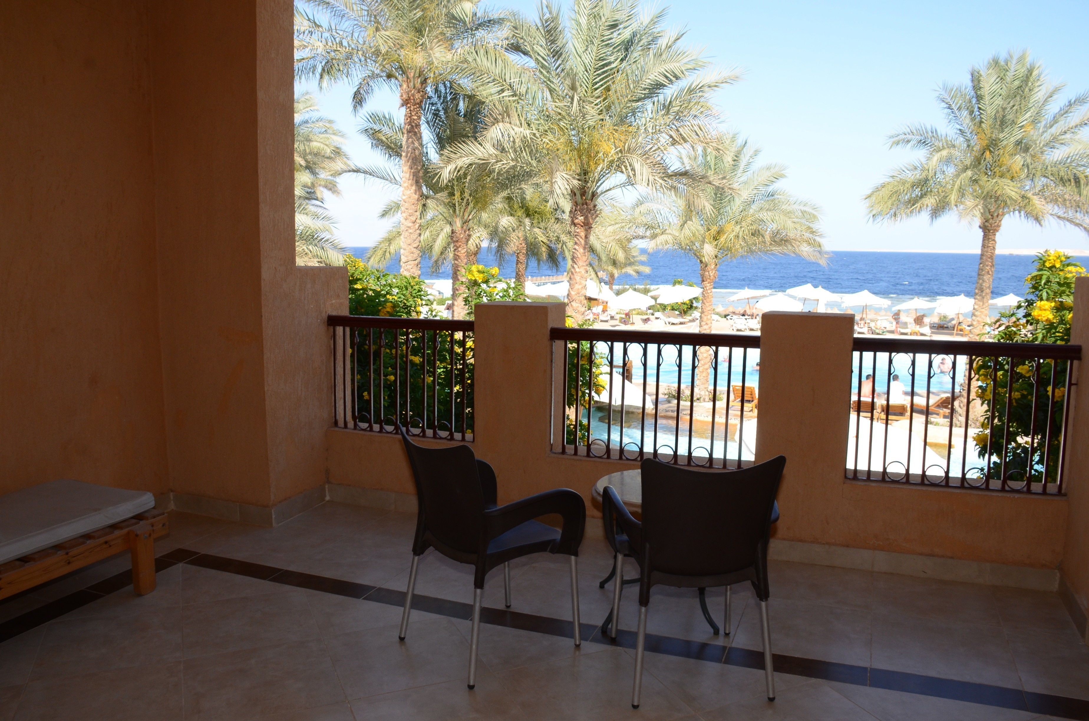 A balcony with two chairs and a bench, overlooking a pool area with palm trees and a view of the sea.