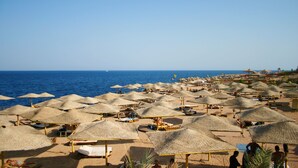 A beach with numerous thatched umbrellas and beach chairs, a clear blue sky, and the ocean in the background. - Falcon Hills Hotel (Sharm El Sheikh)