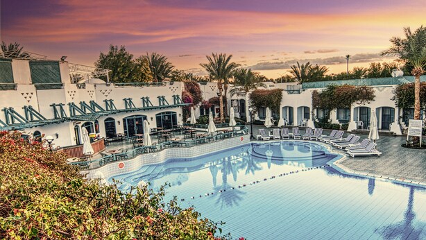 A pool area with lounge chairs and umbrellas, surrounded by buildings with arches and a sunset sky. - Falcon Hills Hotel (Sharm El Sheikh)